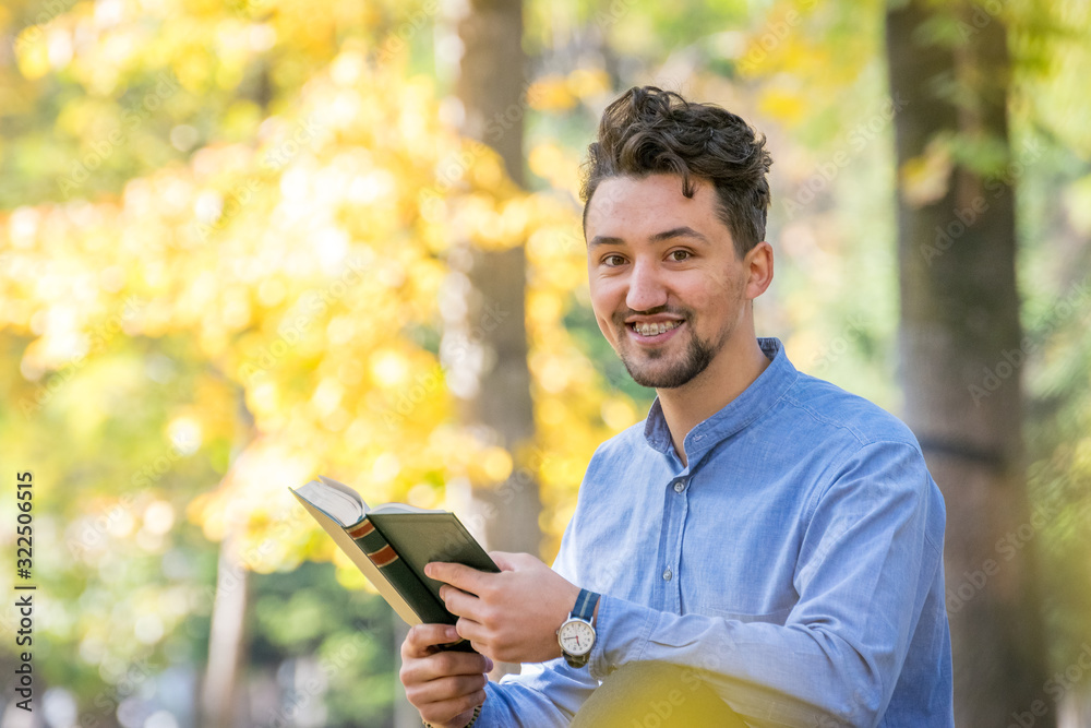 Handsome young man reading a book in a park. Portrait of a young man with a denim jacket and blue shirt reading a book outside. A guy reading a book in a park