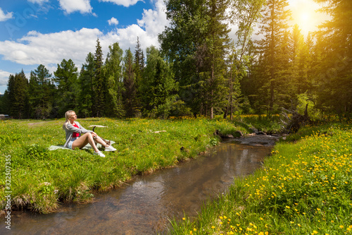 Young beautiful blonde girl in shorts with beautiful legs sits on the banks of a stream and relaxes while listening to the murmur of water on a green edge near the forest on a warm summer day.