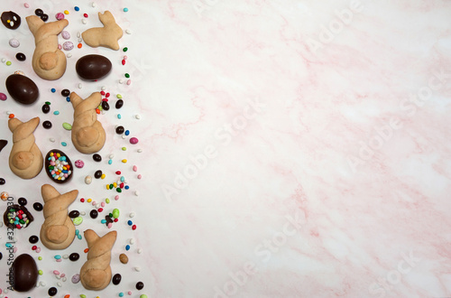 Easter composition. The border is made of homemade buns in the form of Easter bunnies and chocolate eggs of different sizes and colors on a pink marble background. Free space.
