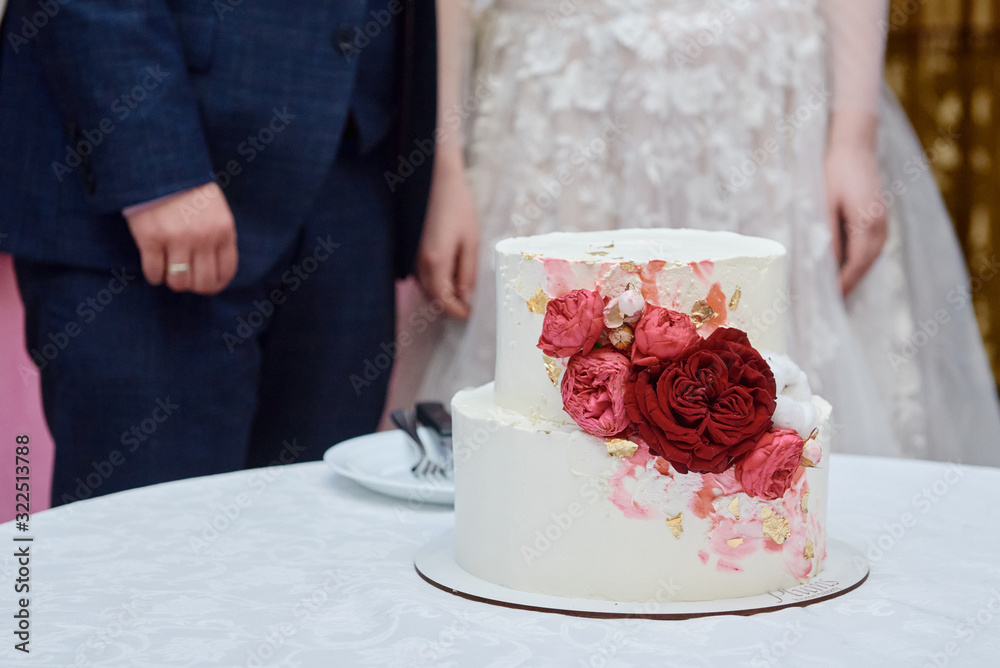 Elegant white wedding cake decorated with pink and red rose flowers on table in restaurant with bride and groom on background, copy space Elegant white wedding cake decorated with pink and red rose flowers on table in restaurant with bride and groom on background, copy space