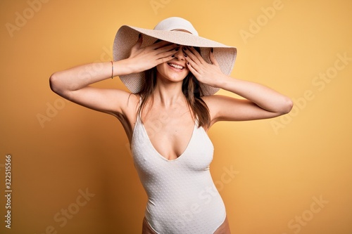 Young beautiful brunette woman on vacation wearing swimsuit and summer hat covering eyes with hands smiling cheerful and funny. Blind concept.