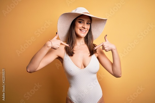 Young beautiful brunette woman on vacation wearing swimsuit and summer hat looking confident with smile on face, pointing oneself with fingers proud and happy.