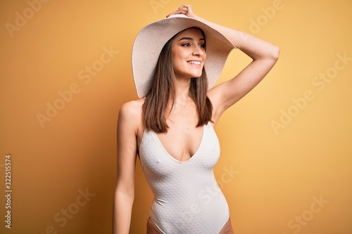 Young beautiful brunette woman on vacation wearing swimsuit and summer hat smiling confident touching hair with hand up gesture, posing attractive and fashionable