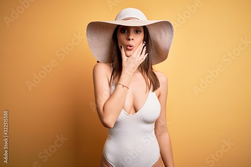 Young beautiful brunette woman on vacation wearing swimsuit and summer hat Looking fascinated with disbelief, surprise and amazed expression with hands on chin