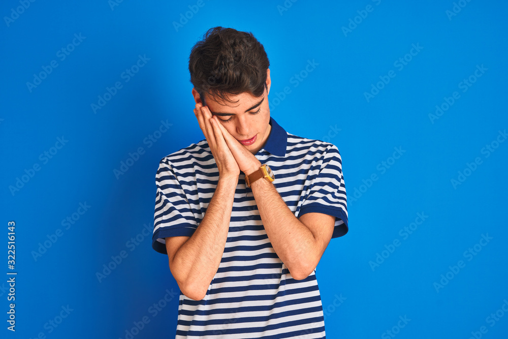 Teenager boy wearing casual t-shirt standing over blue isolated background sleeping tired dreaming and posing with hands together while smiling with closed eyes. Teenager boy wearing casual t-shirt standing over blue isolated background sleeping tired dreaming and posing with hands together while smiling with closed eyes.