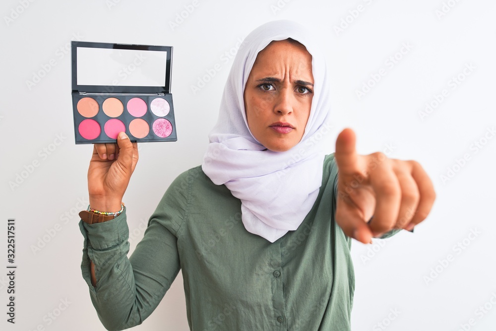 Young Arab woman wearing hijab showing makeup blush palette over isolated background pointing with finger to the camera and to you, hand sign, positive and confident gesture from the front Young Arab woman wearing hijab showing makeup blush palette over isolated background pointing with finger to the camera and to you, hand sign, positive and confident gesture from the front