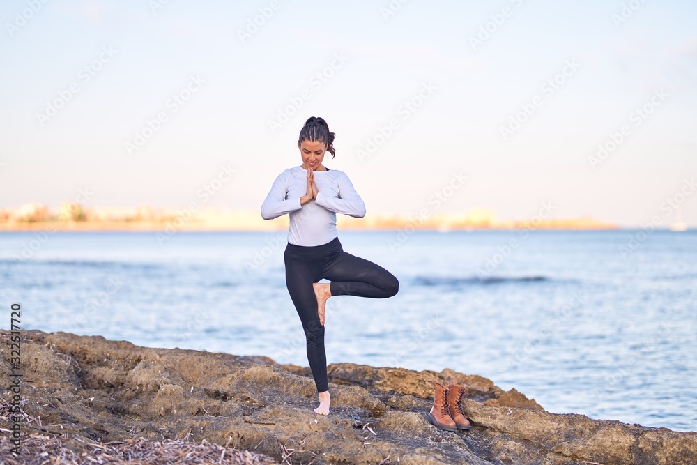 Young beautiful sportwoman smiling happy practicing yoga. Coach with smile on face teaching tree pose at the beach