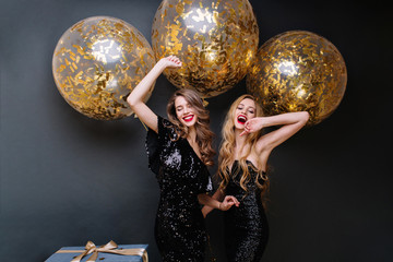 Happy party moments of two fashionable funny young women on black background. Luxury black dress, red lips, long curly hair, brightful mood, having fun, big balloons with golden tinsels