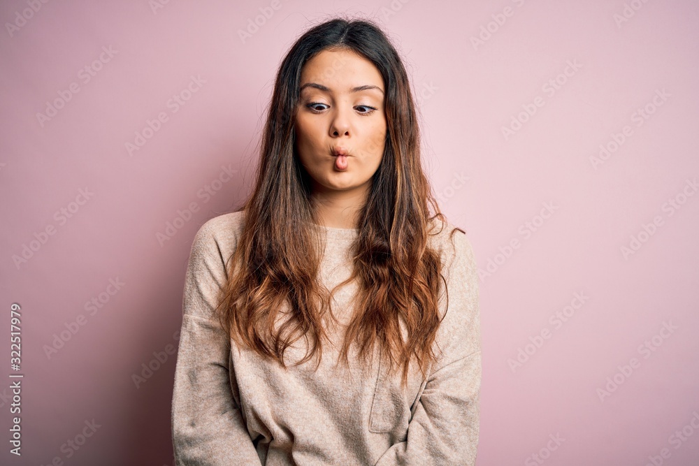 Young beautiful brunette woman wearing casual sweater standing over pink background making fish face with lips, crazy and comical gesture. Funny expression.
