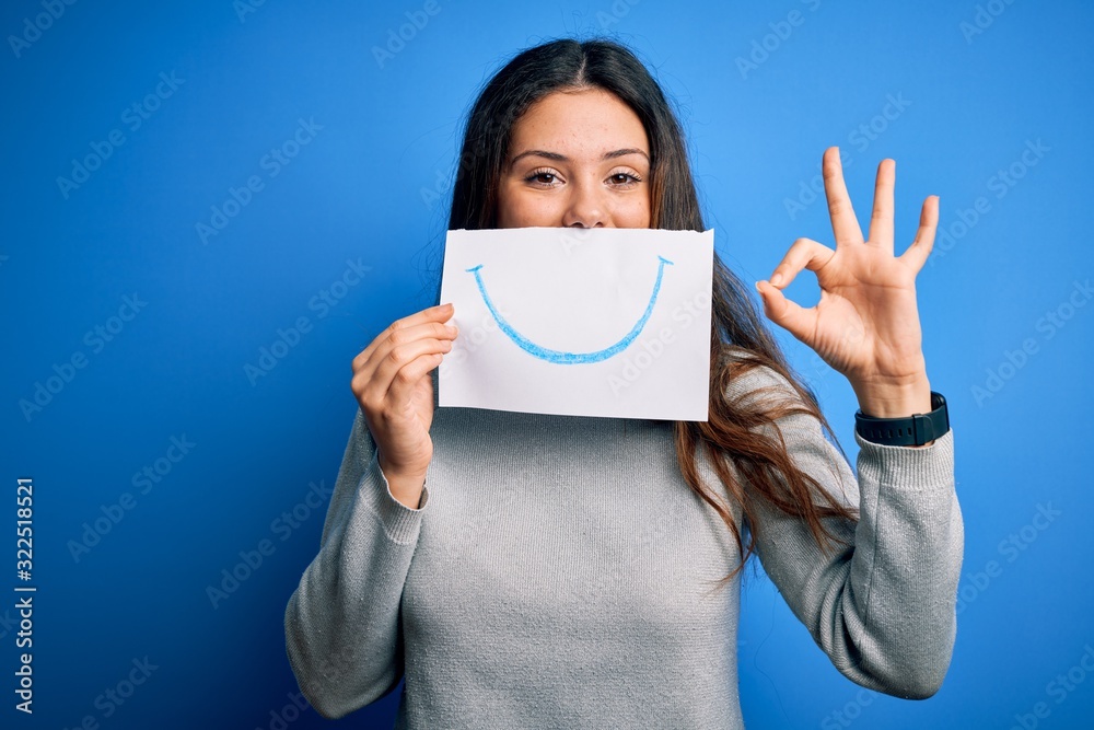 Young beautiful brunette positive woman holding card with smile emotion on mouth doing ok sign with fingers, excellent symbol Young beautiful brunette positive woman holding card with smile emotion on mouth doing ok sign with fingers, excellent symbol