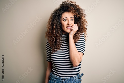 Young beautiful woman with curly hair and piercing wearing casual striped t-shirt looking stressed and nervous with hands on mouth biting nails. Anxiety problem.