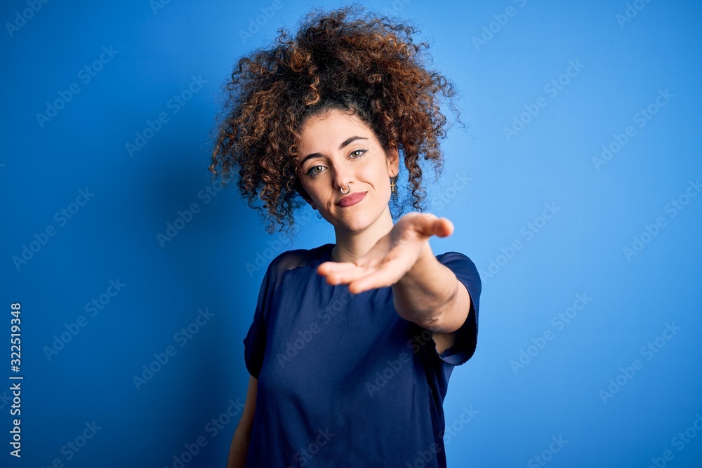 Young beautiful woman with curly hair and piercing wearing casual blue t-shirt smiling friendly offering handshake as greeting and welcoming. Successful business.
