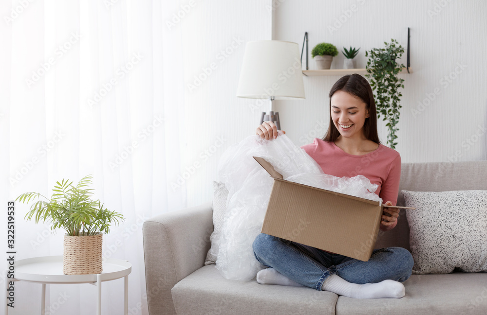 Excited young woman unwrapping parcel, buying goods via internet - obrazy, fototapety, plakaty 