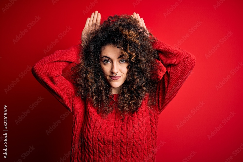 Young beautiful woman with curly hair and piercing wearing casual red sweater Doing bunny ears gesture with hands palms looking cynical and skeptical. Easter rabbit concept. Young beautiful woman with curly hair and piercing wearing casual red sweater Doing bunny ears gesture with hands palms looking cynical and skeptical. Easter rabbit concept.