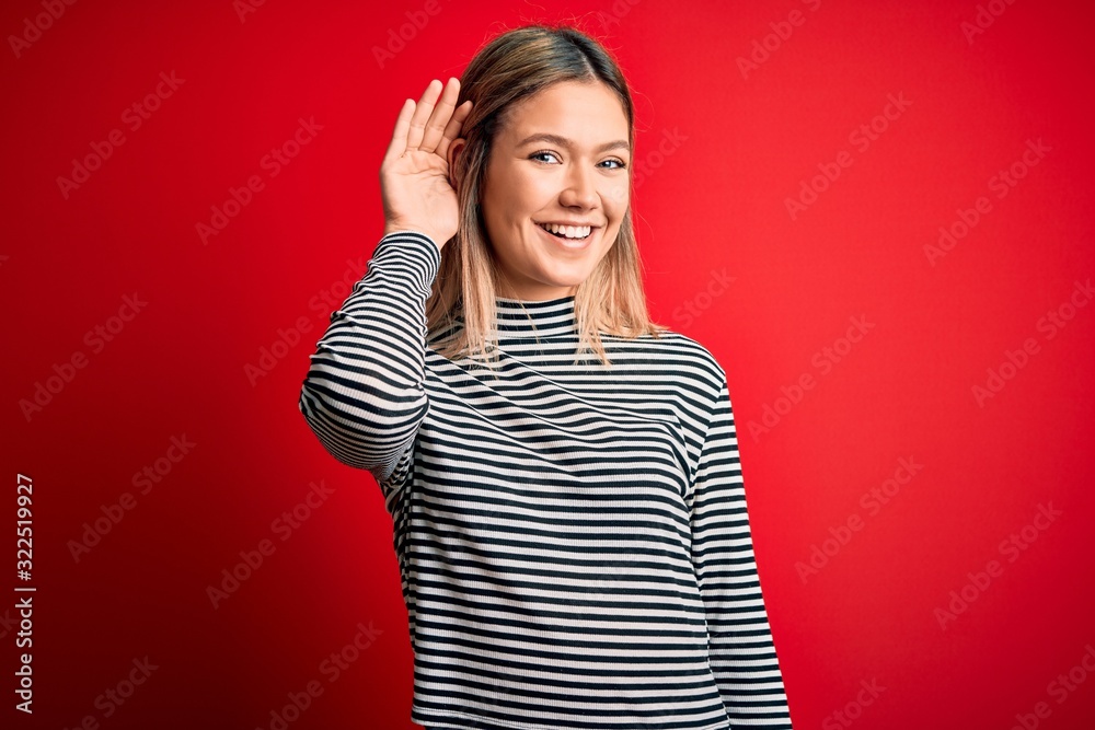 Fototapeta premium Young beautiful blonde woman wearing casual striped sweater over red isolated background smiling with hand over ear listening an hearing to rumor or gossip. Deafness concept.
