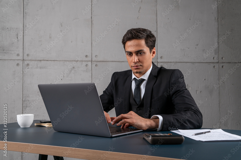 Portrait of serious and concentrated young man in dark suit typing on laptop in loft office. Concept of management and computer science Portrait of serious and concentrated young man in dark suit typing on laptop in loft office. Concept of management and computer science