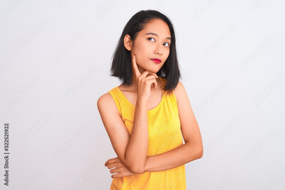 Young chinese woman wearing yellow casual t-shirt standing over isolated white background with hand on chin thinking about question, pensive expression. Smiling with thoughtful face. Doubt concept. Young chinese woman wearing yellow casual t-shirt standing over isolated white background with hand on chin thinking about question, pensive expression. Smiling with thoughtful face. Doubt concept.