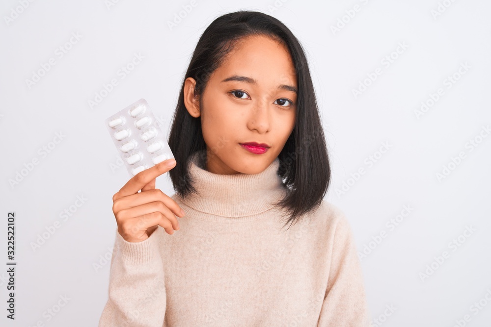 Young beautiful chinese woman holding pills standing over isolated white background with a confident expression on smart face thinking serious Young beautiful chinese woman holding pills standing over isolated white background with a confident expression on smart face thinking serious
