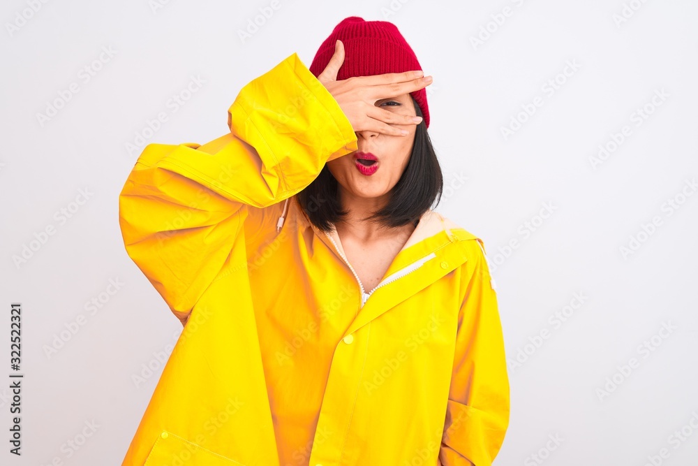 Young beautiful chinese woman wearing raincoat and wool cap over isolated white background peeking in shock covering face and eyes with hand, looking through fingers with embarrassed expression. Young beautiful chinese woman wearing raincoat and wool cap over isolated white background peeking in shock covering face and eyes with hand, looking through fingers with embarrassed expression.