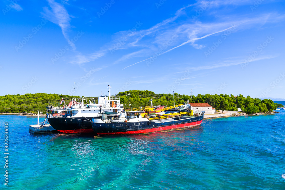 Kornati, Dalmatia. Commercial cargo ship in the port, industrial ships on the sea, transport and logistic concept. Boats in the harbor at the blue lagoon at the island, Adriatic Sea, Croatia.