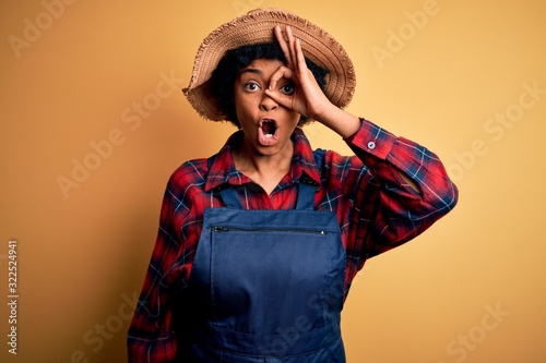 Young African American afro farmer woman with curly hair wearing apron and hat doing ok gesture shocked with surprised face, eye looking through fingers. Unbelieving expression.