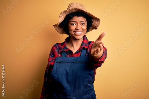 Young African American afro farmer woman with curly hair wearing apron and hat smiling friendly offering handshake as greeting and welcoming. Successful business.