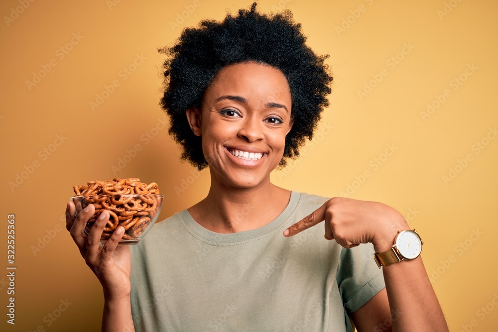 Young African American afro woman with curly hair holding bowl with baked German pretzels with surprise face pointing finger to himself Young African American afro woman with curly hair holding bowl with baked German pretzels with surprise face pointing finger to himself