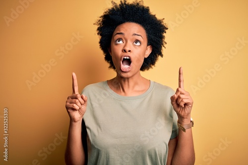 Young beautiful African American afro woman with curly hair wearing casual t-shirt amazed and surprised looking up and pointing with fingers and raised arms.