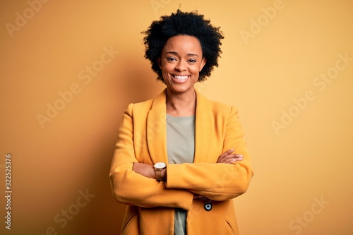 Young beautiful African American afro businesswoman with curly hair wearing yellow jacket happy face smiling with crossed arms looking at the camera. Positive person.