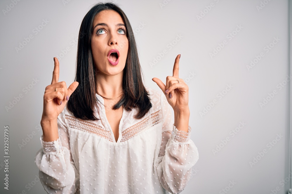 Young brunette woman with blue eyes wearing casual t-shirt over isolated white background amazed and surprised looking up and pointing with fingers and raised arms. Young brunette woman with blue eyes wearing casual t-shirt over isolated white background amazed and surprised looking up and pointing with fingers and raised arms.