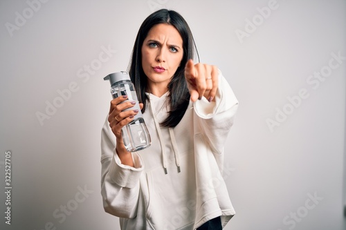 Young brunette sporty woman with blue eyes holding water bottle over white background pointing with finger to the camera and to you, hand sign, positive and confident gesture from the front