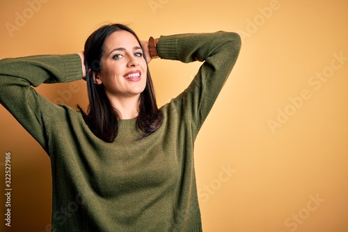 Young brunette woman with blue eyes wearing green casual sweater over yellow background relaxing and stretching, arms and hands behind head and neck smiling happy © Krakenimages.com