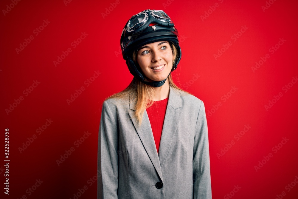 Young beautiful blonde motorcyclist woman wearing motorcycle helmet over red background looking away to side with smile on face, natural expression. Laughing confident. Young beautiful blonde motorcyclist woman wearing motorcycle helmet over red background looking away to side with smile on face, natural expression. Laughing confident.