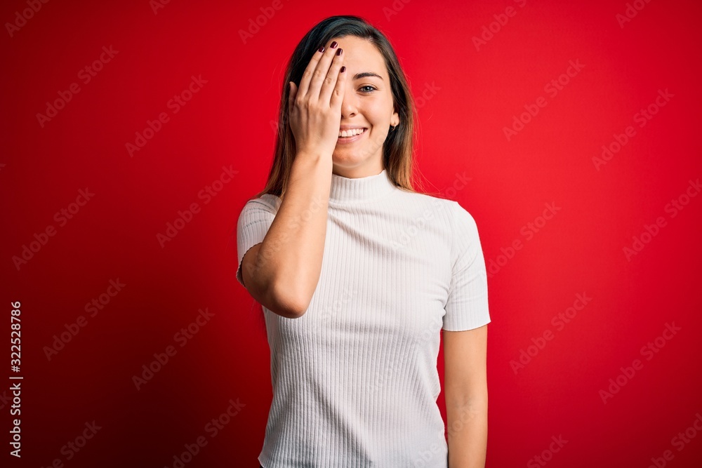 Beautiful blonde woman with blue eyes wearing casual white t-shirt over red background covering one eye with hand, confident smile on face and surprise emotion. Beautiful blonde woman with blue eyes wearing casual white t-shirt over red background covering one eye with hand, confident smile on face and surprise emotion.