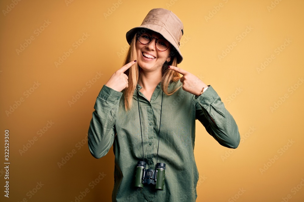 Beautiful blonde explorer woman with blue eyes wearing hat and glasses using binoculars smiling cheerful showing and pointing with fingers teeth and mouth. Dental health concept. Beautiful blonde explorer woman with blue eyes wearing hat and glasses using binoculars smiling cheerful showing and pointing with fingers teeth and mouth. Dental health concept.