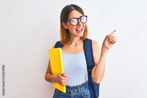 Beautiful redhead student woman wearing a backpack and book very happy pointing with hand and finger to the side