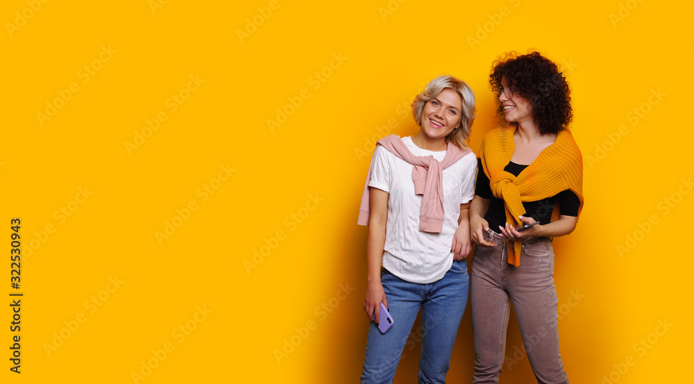 Two smiling and good looking workers are posing on a yellow background holding their phones and sharing their warm emotions Two smiling and good looking workers are posing on a yellow background holding their phones and sharing their warm emotions