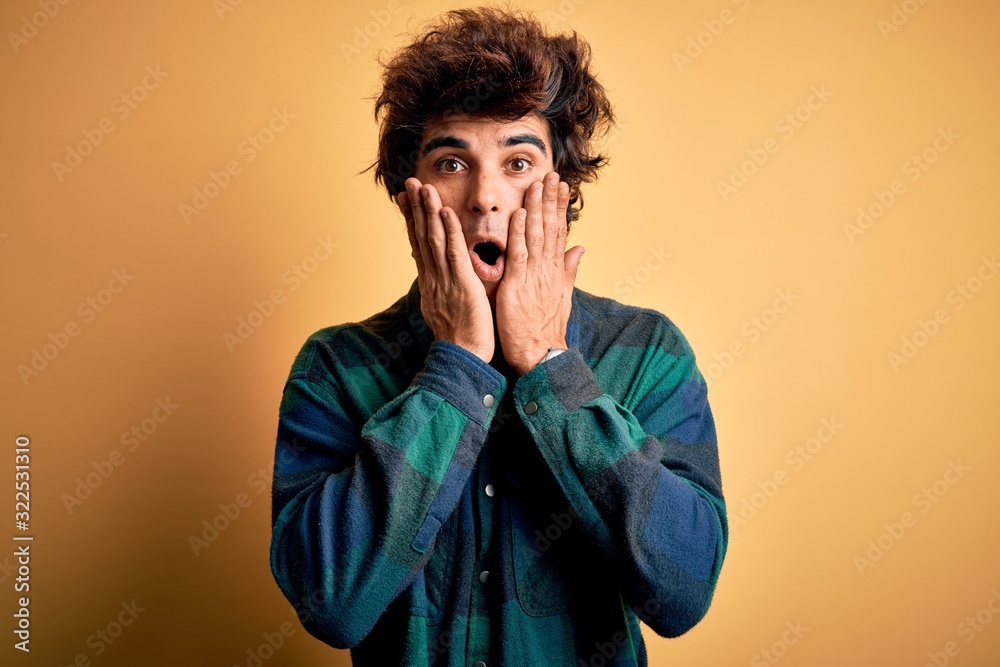 Young handsome man wearing casual shirt standing over isolated yellow background afraid and shocked, surprise and amazed expression with hands on face