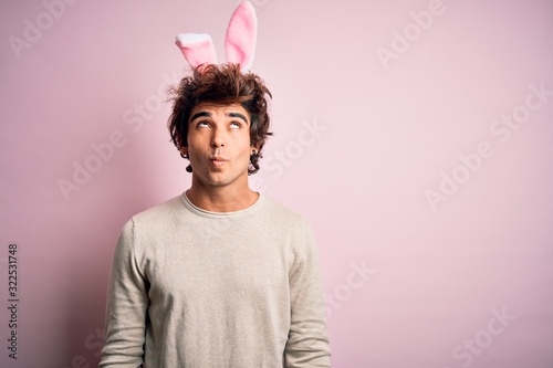 Young handsome man holding easter rabbit ears standing over isolated pink background making fish face with lips, crazy and comical gesture. Funny expression.