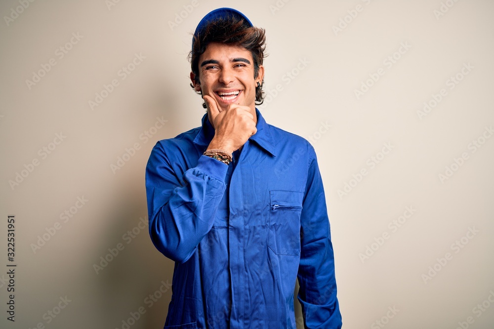 Young mechanic man wearing blue cap and uniform standing over isolated white background looking confident at the camera smiling with crossed arms and hand raised on chin. Thinking positive. Young mechanic man wearing blue cap and uniform standing over isolated white background looking confident at the camera smiling with crossed arms and hand raised on chin. Thinking positive.