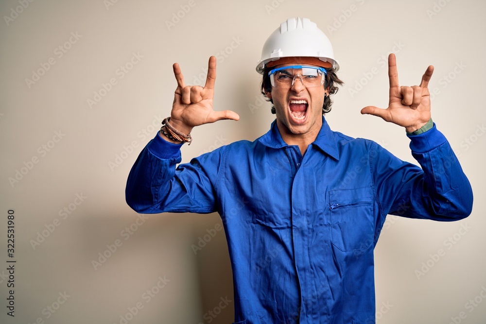 Young constructor man wearing uniform and security helmet over isolated white background shouting with crazy expression doing rock symbol with hands up. Music star. Heavy concept.