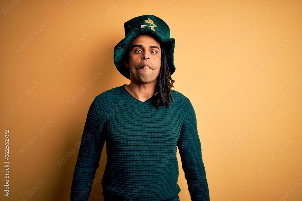 Fototapeta premium Young african american man wearing green hat with clover celebrating saint patricks day making fish face with lips, crazy and comical gesture. Funny expression.