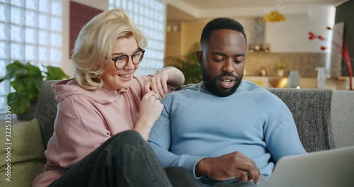 Cheerful mixed ethnicity young couple using notebook for e-commerce in living room, pensive African man and Caucasian woman doing online shopping together watching video movie resting on couch at home