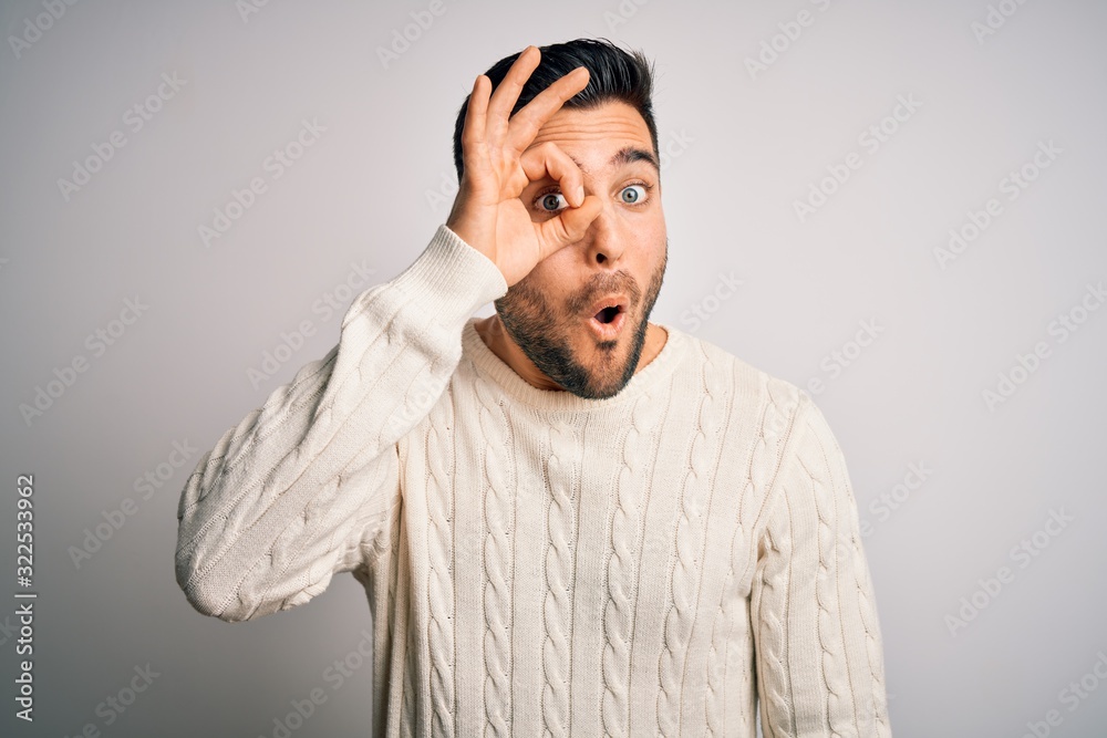 Young handsome man wearing casual sweater standing over isolated white background doing ok gesture shocked with surprised face, eye looking through fingers. Unbelieving expression.