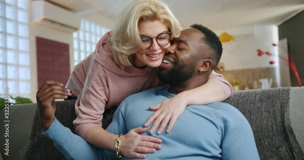 Cute Caucasian woman coming from back to hug and kiss boyfriends sitting on couch at home, pensive man smiling looking at beautiful blonde girlfriend holding mobile phone resting in living room Cute Caucasian woman coming from back to hug and kiss boyfriends sitting on couch at home, pensive man smiling looking at beautiful blonde girlfriend holding mobile phone resting in living room