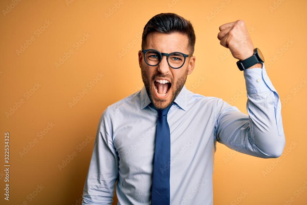 Young handsome businessman wearing tie and glasses standing over yellow background angry and mad raising fist frustrated and furious while shouting with anger. Rage and aggressive concept. Young handsome businessman wearing tie and glasses standing over yellow background angry and mad raising fist frustrated and furious while shouting with anger. Rage and aggressive concept.