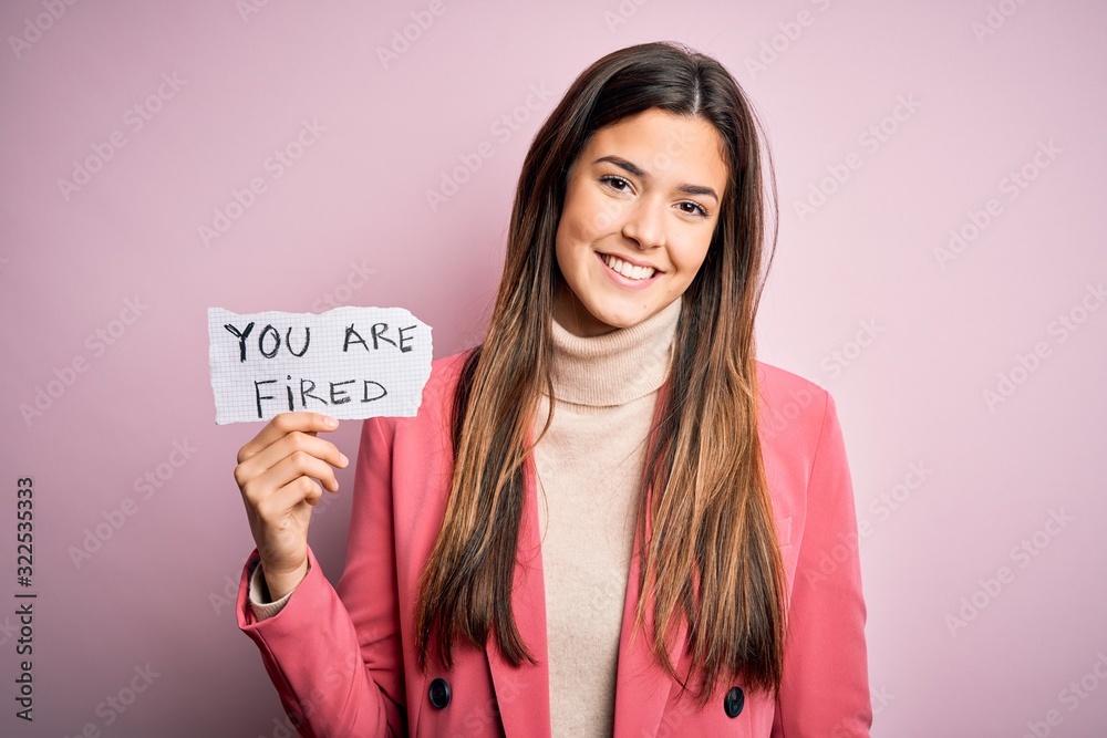 Young beautiful girl holding paper with you are fired message over isolated pink background with a happy face standing and smiling with a confident smile showing teeth Young beautiful girl holding paper with you are fired message over isolated pink background with a happy face standing and smiling with a confident smile showing teeth