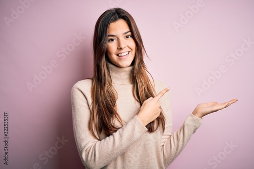 Young beautiful girl wearing casual turtleneck sweater standing over isolated pink background amazed and smiling to the camera while presenting with hand and pointing with finger.