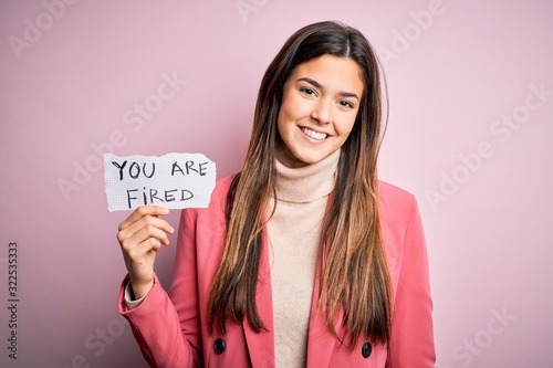 Young beautiful girl holding paper with you are fired message over isolated pink background with a happy face standing and smiling with a confident smile showing teeth