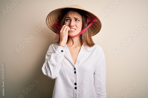 Young beautiful redhead woman wearing asian traditional conical hat over white background looking stressed and nervous with hands on mouth biting nails. Anxiety problem.